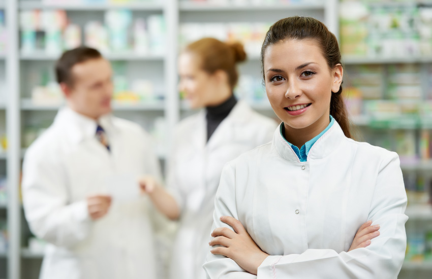 Una farmac&eacute;utica en primer plano con los brazos cruzados sonr&iacute;e mientras dos farmac&eacute;uticos en el fondo charlan